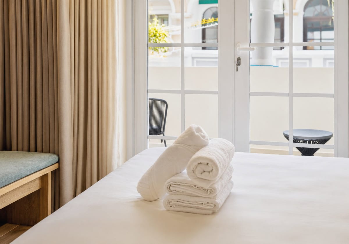 Folded towels on the bed with the balcony doors behind, a woven chair and a dark side table on the balcony