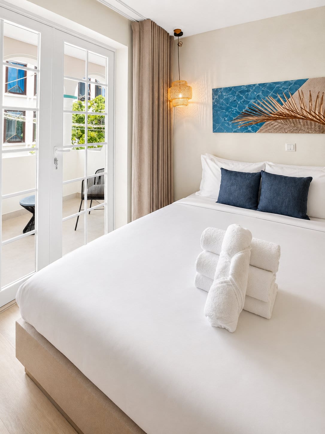 The king bed with folded towels, the balcony doors open beside it and a woven chair on the balcony outside