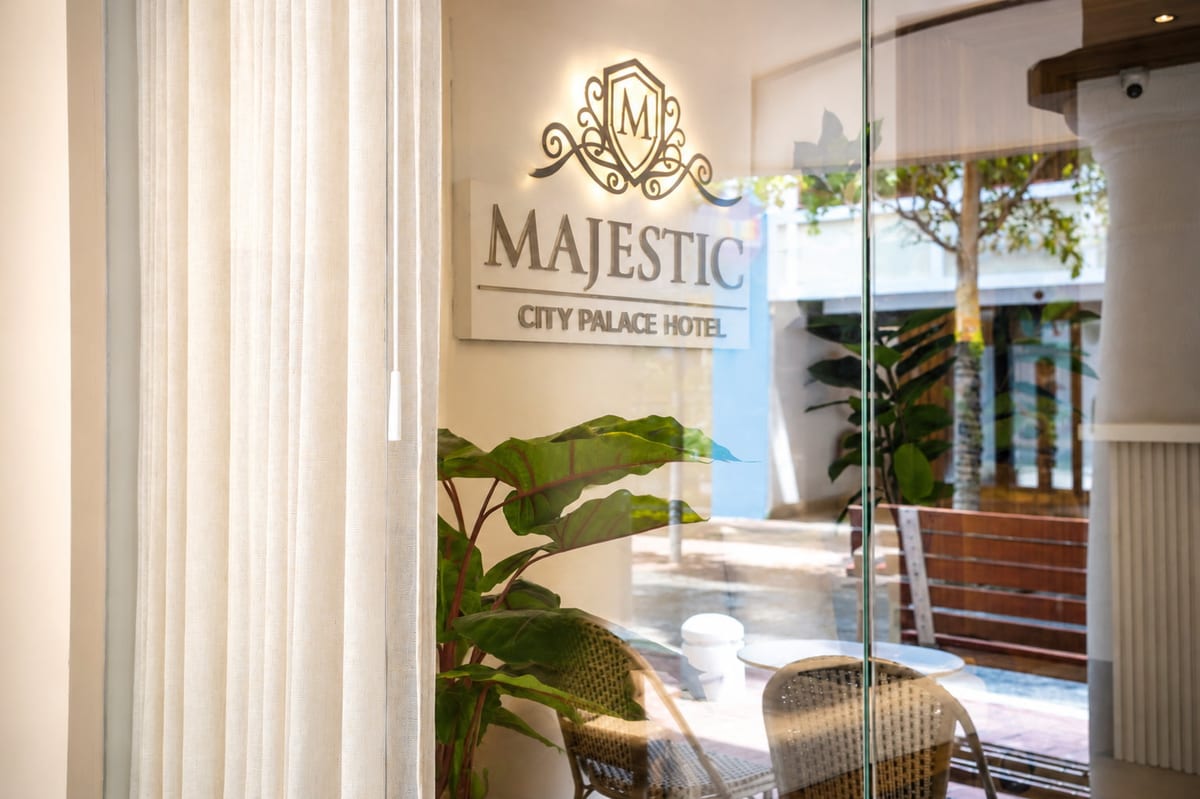 The Majestic City Palace Hotel sign on the lobby wall seen through the glass front, with a linen curtain, a green plant and the rattan terrace chairs of the arcade behind