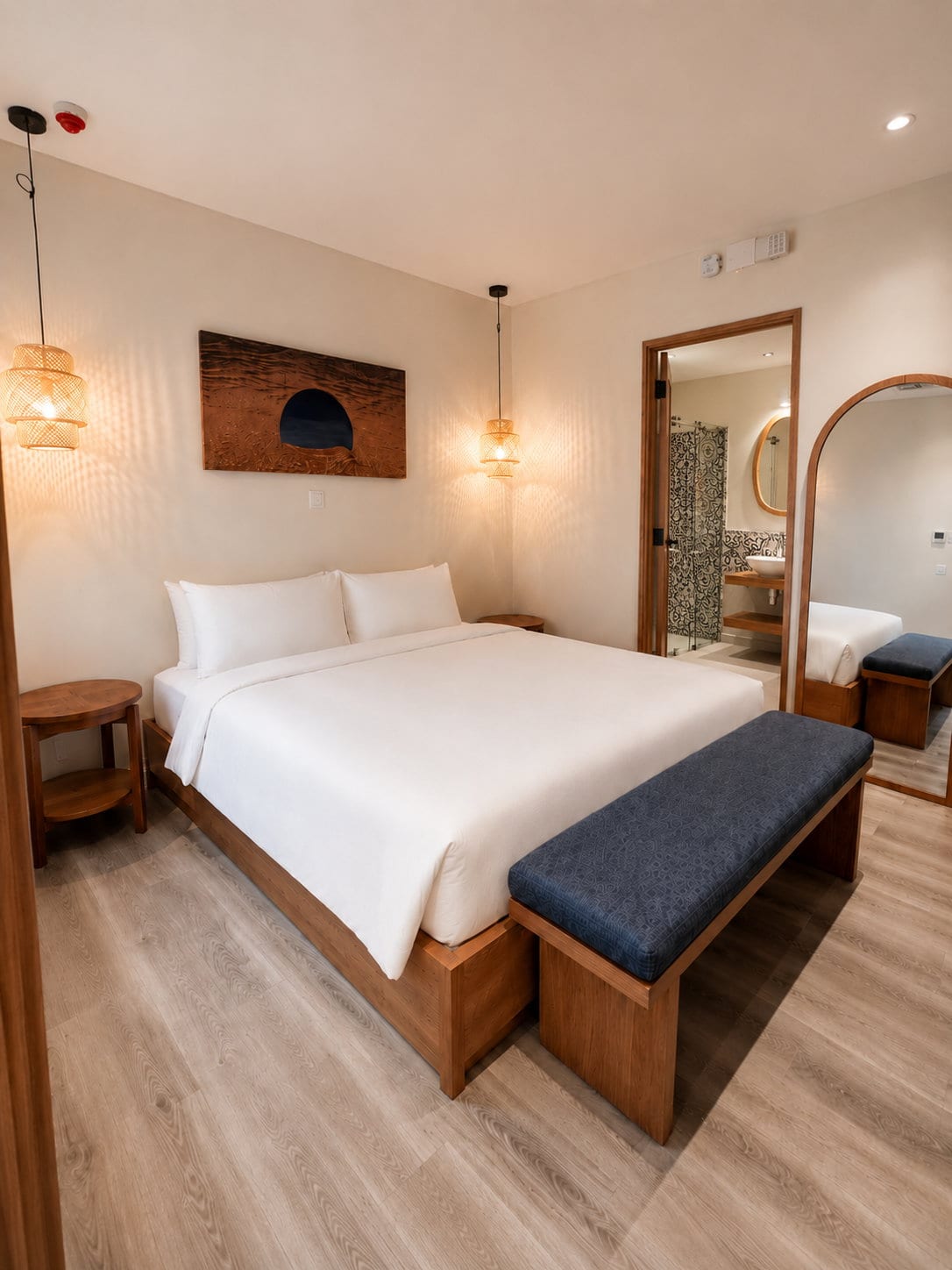 The king bed in white linen with twin rattan pendants, a navy bench at its foot and the tiled bathroom through the open door
