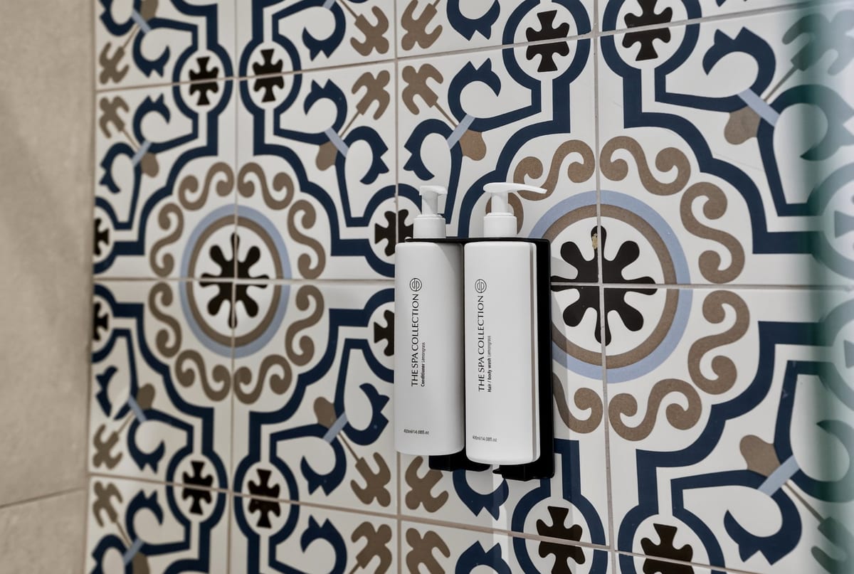 A wall of hand patterned blue, white and ochre majolica tile in a bathroom at Majestic City Palace, with two refillable amenity bottles on a bracket and a brass shower fitting