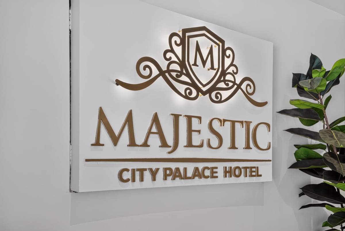 The gold Majestic City Palace Hotel crest and wordmark lit on a plain white plaster wall beside a green fiddle leaf fig