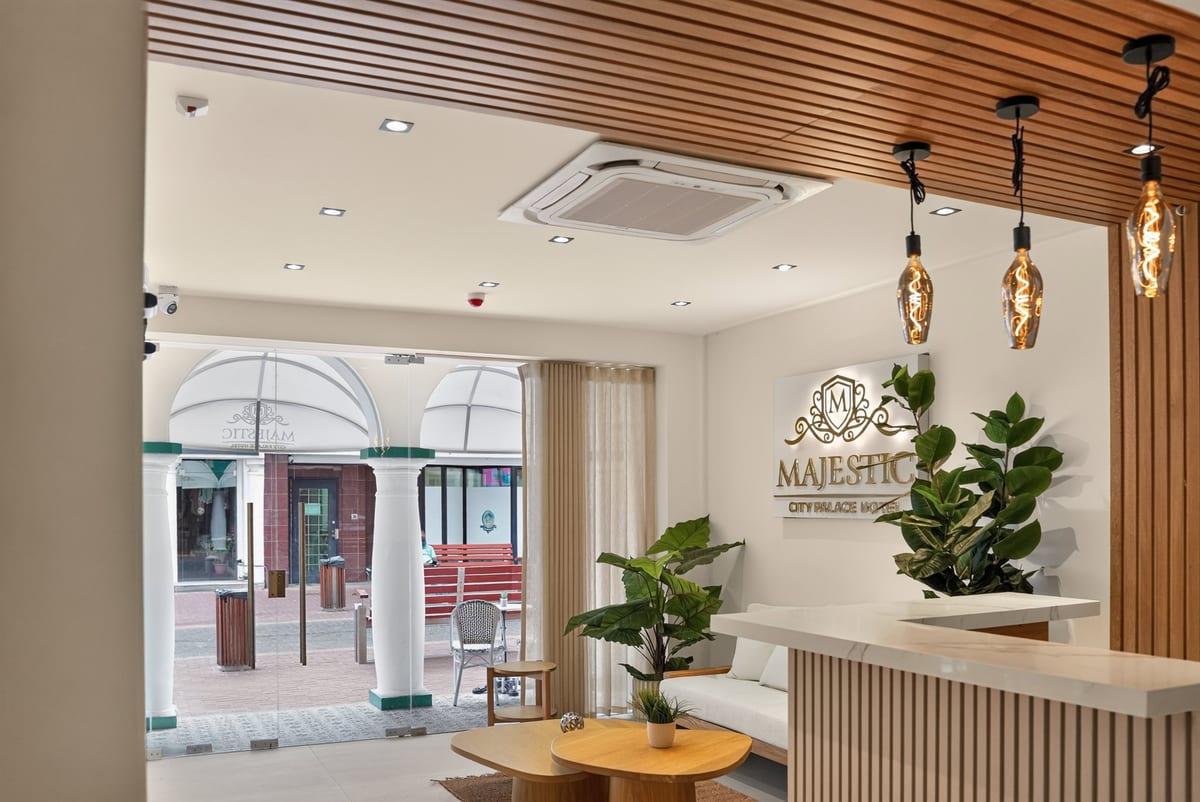 Reception at Majestic City Palace, a fluted desk under a slatted walnut ceiling and filament pendants, the gold crest glowing on the wall and the white arcade beyond the glass front